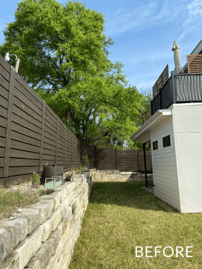Before image of a backyard. There is grass and a stone white wall with a dark brown wooden fence on top of it.