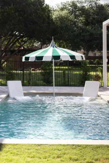 A small outdoor swimming pool in Lakeway features a striped green and white umbrella, two lounge chairs nestled in the water, and a fenced lawn area in the background.
