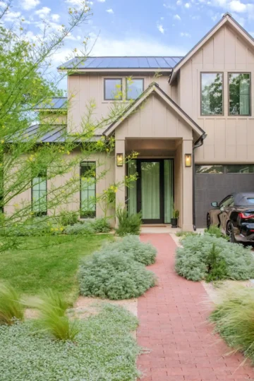 Modern two-story house with tan siding, large windows, and a front entrance. A red brick path leads through landscaped greenery to the front door. A black car is parked in the driveway.