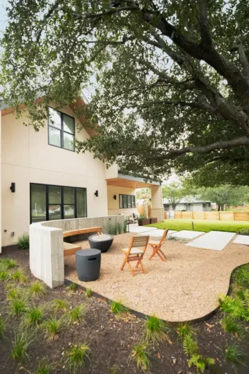 Modern two-story house with large windows, a gravel patio area, wooden chairs, and a fire pit under a large tree in a landscaped yard.