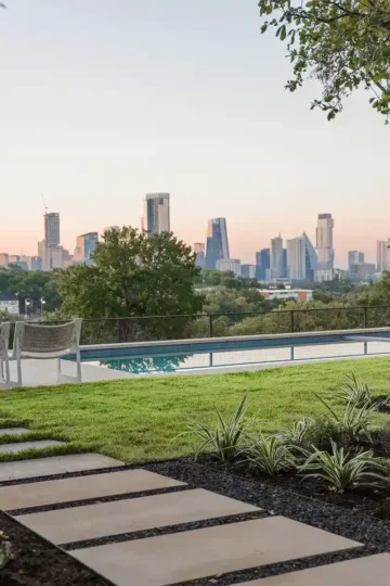 A landscaped backyard with a stone path, lounge chairs, pool, and a view of a city skyline at sunset.