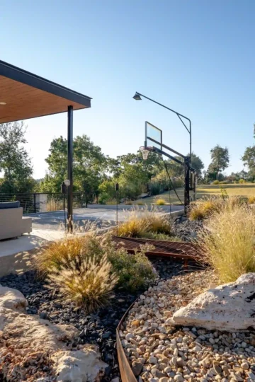 Outdoor basketball court next to a modern patio area, with surrounding landscaping featuring ornamental grasses, rocks, and trees under a clear sky.