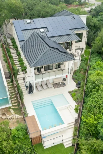 A modern two-story house near Mount Bonnell with solar panels, a terrace, and a backyard featuring a rectangular pool and hot tub, surrounded by greenery and adjacent properties.