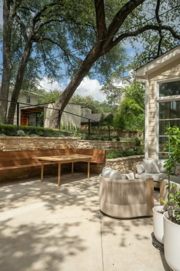 A stone patio with a wooden bench, a round cushioned chair, a rectangular table, potted plants, and large trees providing shade in a residential backyard.