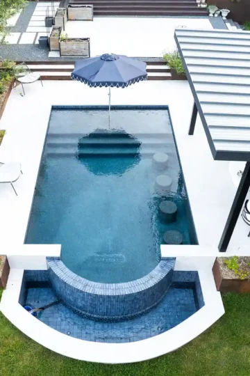 A rectangular backyard pool with a built-in hot tub, shaded seating area, umbrella, chairs, and surrounding green grass and landscaping.